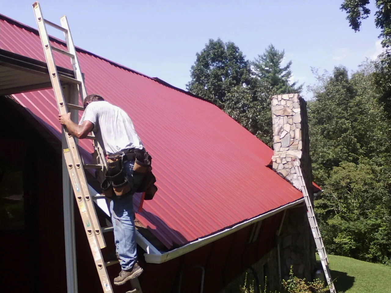 Roofer on a ladder beside a red metal roof and stone chimney — Soffit & Fascia Repair by Roofing Brothers in Saint Charles